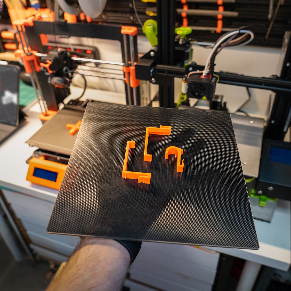 A hand holding a print bed with two orange 3D-printed wall hooks